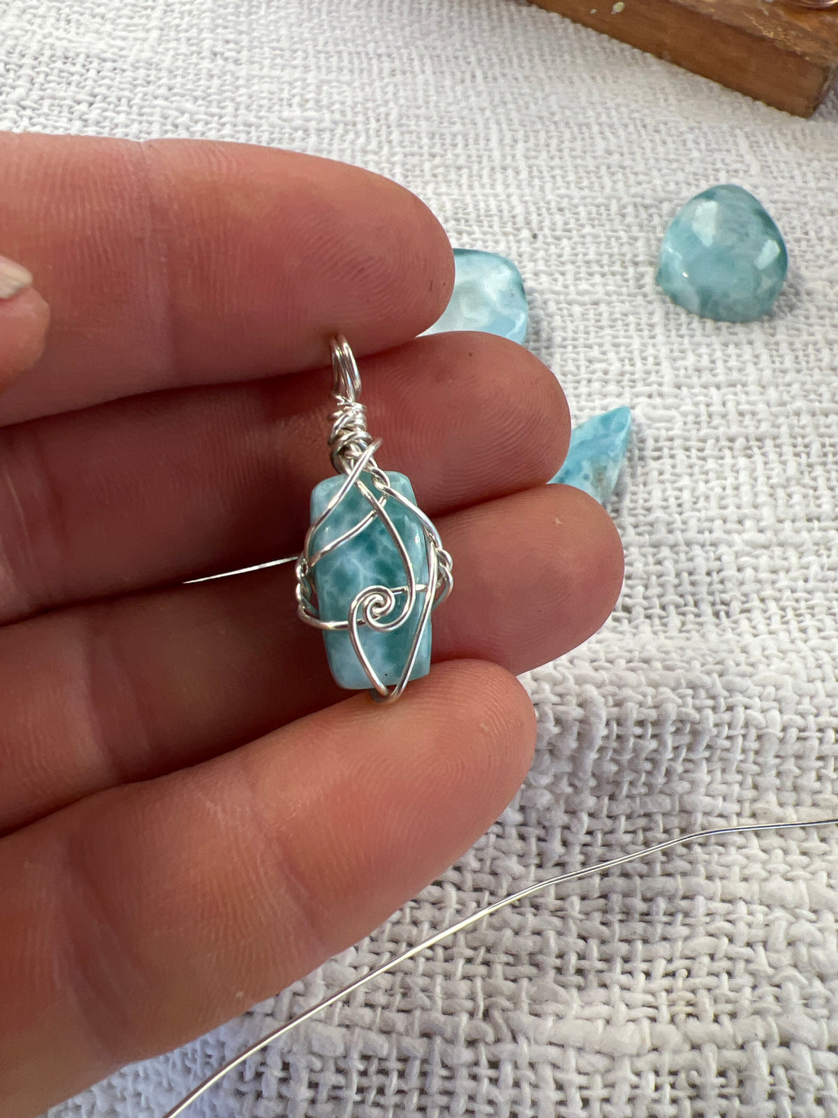 Small handmade Larimar pendant wire-wrapped in sterling silver, held in fingers with loose Larimar stones on linen